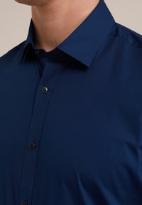 Camisa de botones azul marino con cuello puntiagudo, textura suave y botones negros. Vista en primer plano del frente.