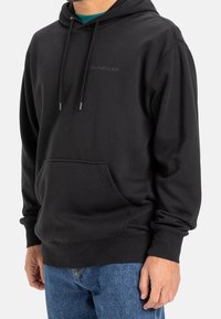 Uomo che indossa una felpa nera con cappuccio e tasca frontale, insieme a jeans blu, in piedi contro uno sfondo chiaro e semplice.