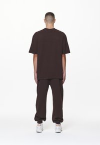 Camiseta oversized marrón de manga corta combinada con pantalones de chándal marrones a juego. El modelo lleva zapatillas blancas, posando frente a un fondo blanco liso.