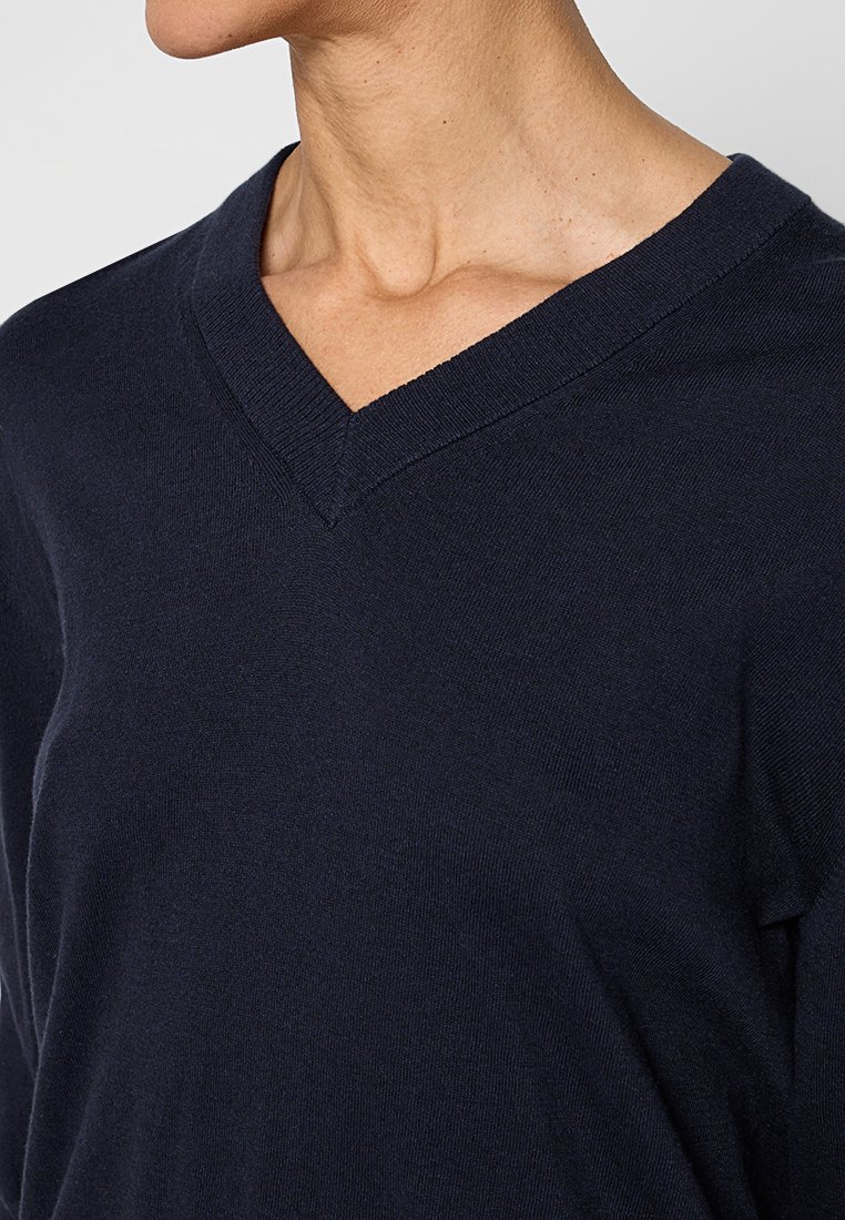 Person, der einen dunkelblauen V-Ausschnitt-Pullover trägt, zeigt Oberkörper und Hals in einer neutralen Pose vor einem schlichten Hintergrund.