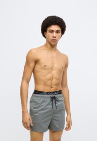 Szürke fürdőnadrág elasztikus fekete derékpánttal, amelyen a márkanév szerepel. Könnyű anyagból, térdhosszúságú, megkötőzsinórral.