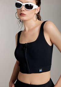 Top corto a coste nero con chiusura a zip frontale, spalline larghe e un patch con logo sull'orlo. Il modello indossa occhiali da sole bianchi e orecchini a cerchio.