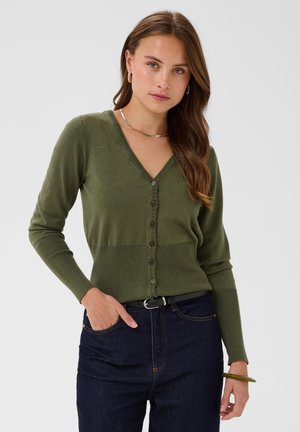 Femme aux longs cheveux bruns portant un cardigan vert olive à col en V, un jean foncé, des créoles et un collier, debout avec une main dans la poche.
