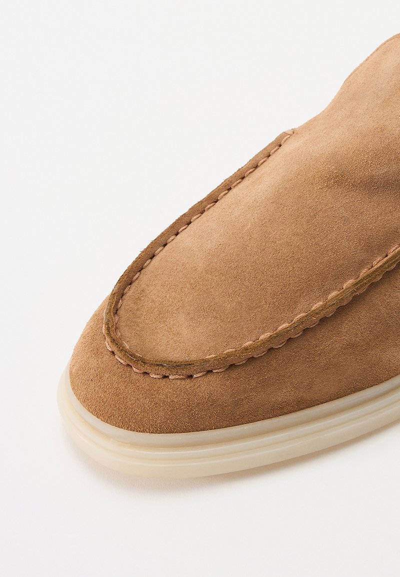 Mocassin en suede marron avec un bout arrondi, des coutures contrastantes le long du bord, et une semelle en caoutchouc de couleur claire.