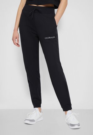 Woman standing wearing black Calvin Klein joggers with drawstring waist and pockets, paired with white sneakers with silver details.