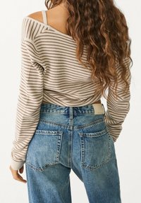 Suéter a rayas beige y marrón con diseño de hombros al aire, combinado con jeans de mezclilla azul de cintura alta que tienen dos bolsillos traseros.