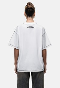 Wit oversized t-shirt met een ronde halslijn, korte mouwen en contrasterende stiksels. De achterkant toont een logo in zwarte tekst.
