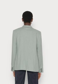 Blazer verde-claro com um corte descontraído, lapelas notched e punhos abotoados, combinado com calças escuras com riscas. Textura de tecido suave.