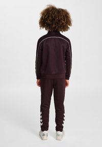 Bordowy dres z białymi wstawkami, w skład którego wchodzi kurtka z zamkiem i zwężane spodnie z akcentami w kształcie zygzaka po bokach. Noszony z jasnymi sneakersami.