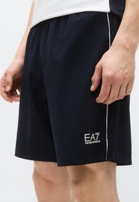 Tmavomodré športové šortky z ľahkej tkaniny; majú bielu značku a bočné pruhy; elastický pás zabezpečuje pohodlné nosenie.