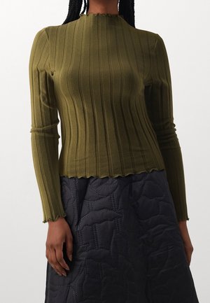 Mujer que lleva una blusa de manga larga de canalé en color verde oliva con bordes en forma de onda y una falda negra acolchada con un patrón de costura floral.