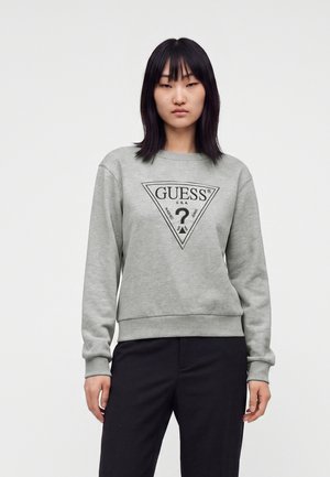 Жена с права черна коса, носеща сива суитшърт Guess и черен панталон, стояща пред светъл фон.