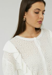 Cardigan tricoté blanc avec un motif en losange, présentant des manches volantes et des boutons en perles sur le devant. Texture lisse, design léger.