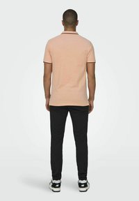 Camisa polo en melocotón claro con ribete negro en las mangas y el cuello, combinada con pantalones negros y zapatillas en blanco y negro. Vista desde la parte posterior.