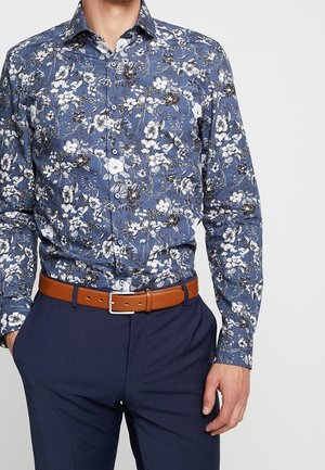Homme portant une chemise à motifs floraux bleue, une ceinture en cuir marron, et un pantalon bleu marine, les mains dans les poches.