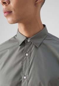 Camisa de botón gris con cuello standard, que presenta una textura suave y botones blancos a lo largo de la tapeta. Vista en primer plano del escote.