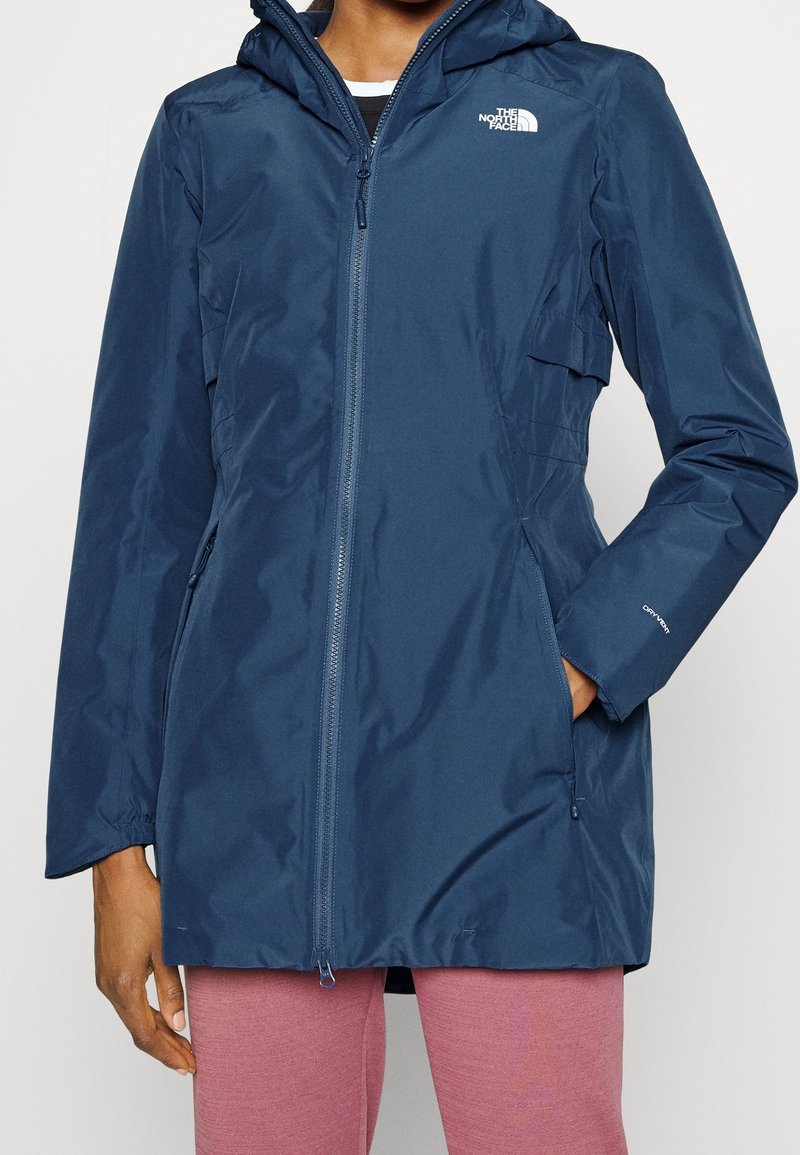 Femme portant une veste à capuche zippée bleu marine North Face, les mains dans les poches et un pantalon rose sur fond uni.