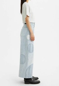 Jupe en denim bleu clair avec des motifs circulaires de tailles variées. Taille haute, poches latérales, associée à un t-shirt gris et des bottes noires.