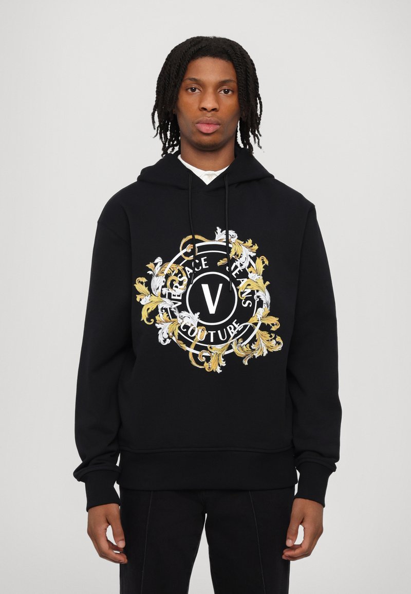 Juodas gobtuvas su centriniu logotipu, puoštu auksinėmis ir baltomis gėlėmis bei užrašu "Versace Jeans Couture."