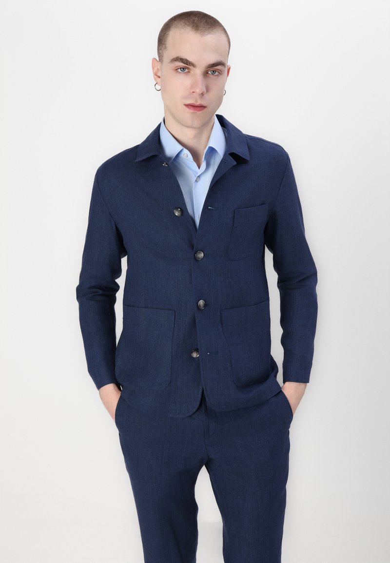 Marineblaue Leinen-Sakkokjacke mit entspannter Passform, Reverskragen, sechs Knöpfen und zwei Fronttaschen; kombiniert mit einem hellblauen Hemd.