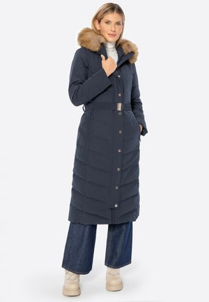 Femme portant un long manteau matelassé bleu marine avec col en fourrure, un pull à col montant, un jean à jambes larges et des bottes beiges, debout, une main dans la poche.