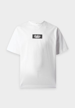 Homme portant un t-shirt blanc avec un logo noir et un jean ample bleu clair, debout avec une main dans la poche et des baskets noires.