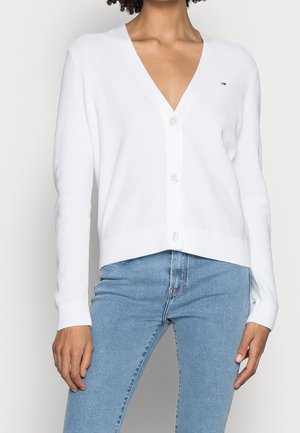 Femme portant un cardigan blanc à boutons et col en V, associé à un jean bleu clair, debout devant un fond clair uni.