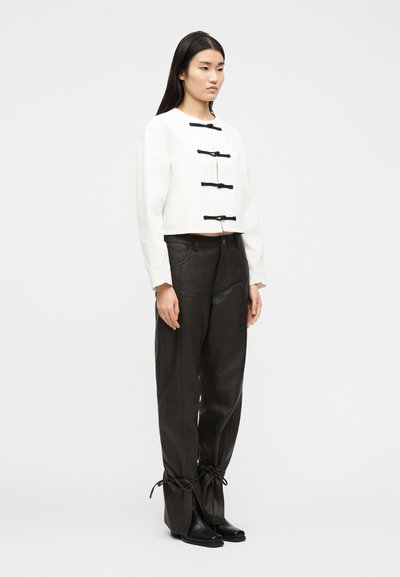 Γυναίκα που στέκεται μπροστά σε λευκό φόντο, φορώντας λευκό cropped τοπ με μακριά μανίκια και μαύρα κουμπώματα, και άνετο μαύρο παντελόνι με δεμένα τελειώματα και μαύρες μπότες.