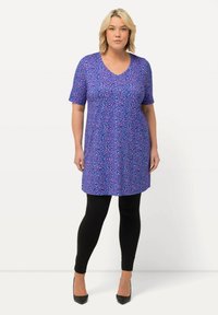 Túnica floral morada con mangas cortas, escote en V y un corte holgado, combinada con leggins negros y tacones negros. Textura de tela suave.