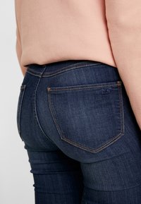 Mörka denimjeans med en figurnära design, med en bakficka med en liten logo och kontrasterande orange sömmar längs sömmarna.