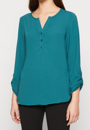 Blouse teal à manches longues en tissu léger, présentant un décolleté en V, trois boutons sur le devant et des poignets retroussés.
