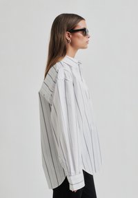 White striped shirt with a high collar, featuring wide sleeves and vertical navy stripes. Model wears sunglasses, shown in profile.