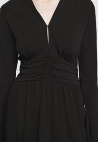 Robe noire à manches longues avec taille froncée et décolleté en V, montrant le haut du torse et la partie médiane du porteur.