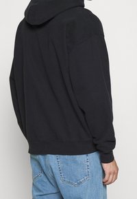 Černá mikina s kapucí a volným střihem, klokáním kapsou, žebrovanými manžetami a lemem, vyrobená z měkké bavlněné směsi. Nahoře světle modré džíny.