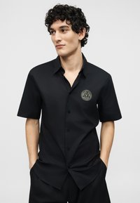 Νεαρός με σγουρά μαλλιά, φοράει μαύρο πουκάμισο με κοντά μανίκια και κουμπιά, με κυκλικό λογότυπο Versace Jeans Couture στο στήθος.