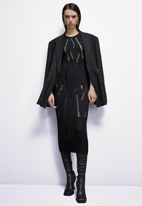 Abito nero lavorato a maglia con dettagli cut-out, indossato sotto un blazer nero oversized. Abbinato a stivali in pelle nera al ginocchio e orecchini vistosi.