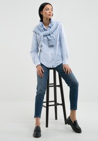 Camicia azzurro chiaro con bottoni e jeans scuri; un maglione blu drappeggiato sulle spalle; mocassini neri; seduto su uno sgabello alto e scuro.