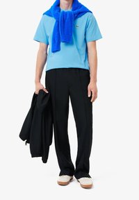 Světle modré tričko krátkého rukávu, tmavě černé kalhoty, modrý svetr uvázaný kolem krku, bílé boty s hnědými podrážkami, černá bunda v ruce.