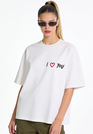 Femme avec queue de cheval haute portant des lunettes de soleil noires, un t-shirt blanc oversized avec le texte "I heart you" et un pantalon vert olive.