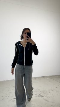 Fekete cipzáras kapucnis pulóver, fehér zsinórral, fehér felsőre húzva, világosszürke bővített lábú farmerrel párosítva. Egyszerű, laza szett.