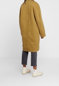 Abrigo largo caqui hecho de una tela texturizada, combinado con pantalones grises recortados y zapatillas altas color crema con acentos azules.