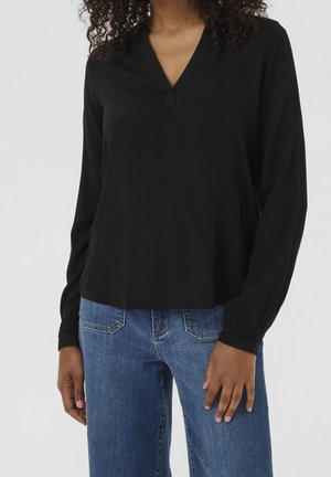 Vrouw die een zwarte blouse met lange mouwen en V-hals draagt, en een blauwe spijkerbroek met zakken, staand tegen een effen achtergrond.