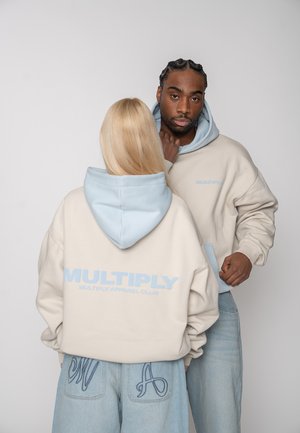 To modeller iført beige hættetrøjer med lyseblå hætte og teksten "MULTIPLY", kombineret med lyseblå denimjeans mod en ensfarvet baggrund.