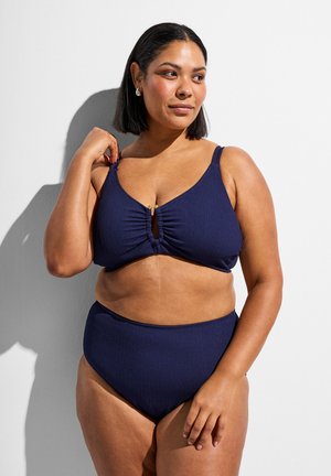 Kobieta o włosach do ramion, ubrana w granatowy prążkowany bikini z marszczonym topem i majtkami z wysokim stanem, stojąca na białym tle.