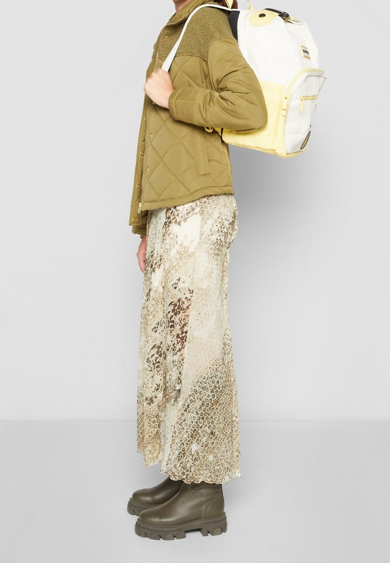 En person står på sidan, iklädd en grön vadderad jacka, en lätt mönstrad kjol och gröna stövlar, medan hen bär en vit och gul ryggsäck.