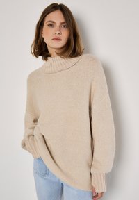 Femme aux cheveux bruns mi-longs portant un pull col roulé beige et un jeans bleu clair, se tenant contre un mur blanc.