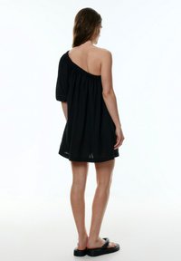 Zwarte one-shoulder jurk met een losse fit, elastische halslijn en korte pofmouw. Soepele stof en hoogte boven de knie. Zwarte sandalen.