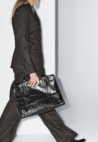 Sac en cuir noir gaufré façon crocodile avec double poignée, forme structurée et finition brillante, tenu par une personne portant un costume rayé foncé.