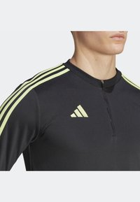 Black athletic zip-up top featuring three yellow accent stripes on each shoulder and an Adidas logo on the chest. Smooth, fitted design.