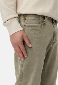 Beżowy sweatshirt z ściągaczami na rękawach w połączeniu z oliwkowymi dżinsami, które mają przednią kieszeń, widoczny guzik oraz subtelną teksturę.